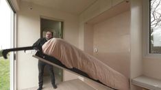 a man standing next to a bed on top of a wooden floor in a room