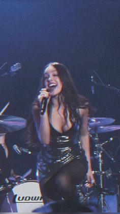 a woman singing into a microphone while sitting in front of drums and drum sticks on stage