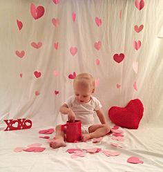 a baby sitting on the floor playing with a red cup and pink heart shaped confetti