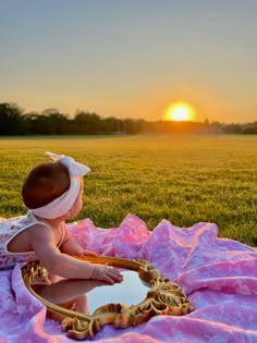 a baby sitting on a blanket in the middle of a field looking at a mirror