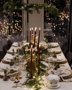 a table set for christmas dinner with candles and greenery on the table, surrounded by festive lights