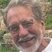 an older man with white hair and beard smiling at the camera while standing in front of some bushes