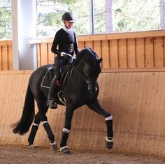 a woman riding on the back of a black horse in an indoor arena with wooden walls