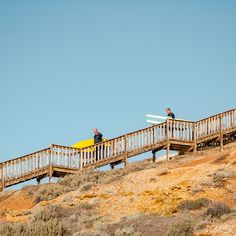 two people with surfboards are standing on a wooden bridge over dry grass and bushes