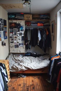 an unmade bed in a small room with clothes hanging on the wall and closet doors open