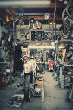 two men are sitting on the top of a motorcycle in a garage filled with tools