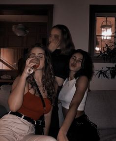 three young women sitting on a couch drinking from bottles in the dark, with one woman standing behind them