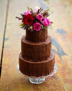 a three layer chocolate cake with flowers on top sitting on a wooden table next to a glass plate