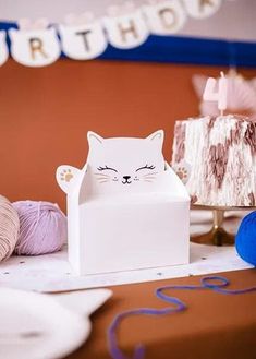 a white cat sitting in a box on top of a table next to balls of yarn