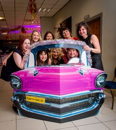 a group of women posing in front of a pink car with the hood up and windows down