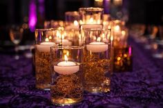 many lit candles are in glass vases on a table with purple cloth and flowers