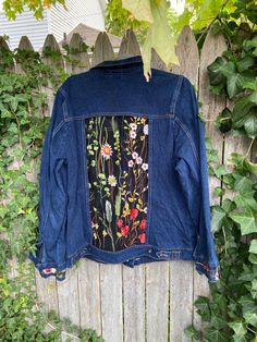 a denim jacket with flowers on it hanging from a wooden fence next to green leaves