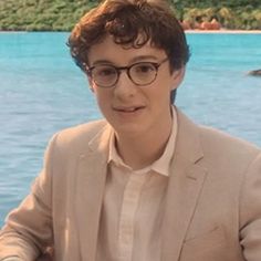 a young man wearing glasses sitting in front of the ocean with an island behind him