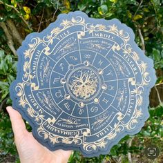 a hand holding up a blue and gold wheel of the year coaster in front of some trees