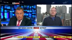 two men are sitting down talking to each other on the television set in front of city buildings