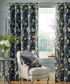 a living room with floral curtains and a chair in front of the window, along with a vase filled with flowers