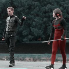 two people dressed in black and red are standing on the street with swords behind them