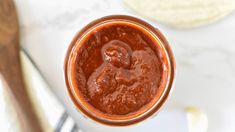 a jar filled with red sauce sitting on top of a white counter next to a wooden spoon