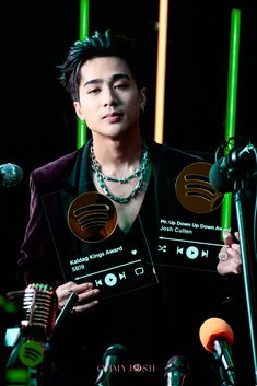 a man holding up two awards in front of microphones with green lights behind him