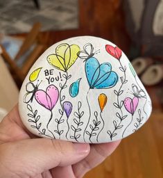 a hand holding a painted rock with flowers and hearts on it that says be you