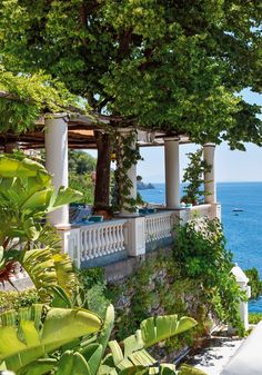 an outdoor dining area overlooks the ocean and is surrounded by greenery, with white pillars