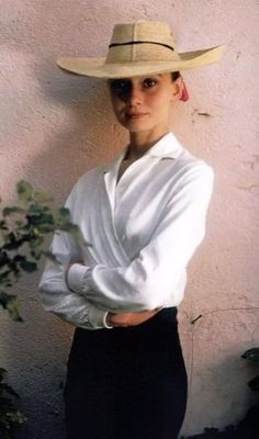 a woman in a white shirt and hat standing next to a wall with her arms crossed