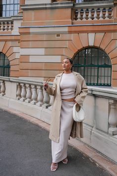 a woman standing on the street in front of a building wearing a tan coat and white skirt