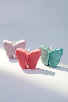 three butterfly shaped candles sitting next to each other on a white surface with light coming from behind them