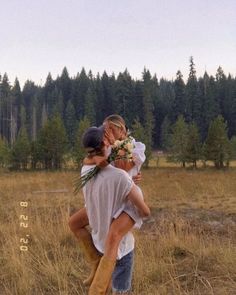 a man carrying a woman in the middle of a field with pine trees behind him