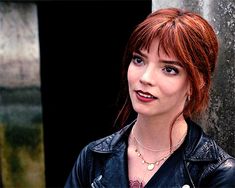 a woman with red hair wearing a black leather jacket and necklace standing in front of a wall