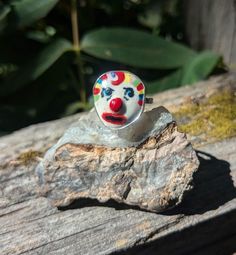 a clown face ring sitting on top of a piece of wood next to a rock