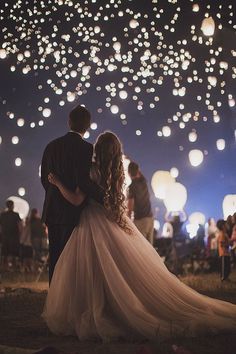 a bride and groom standing in front of the sky full of lanterns at night time