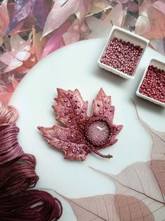 two white bowls filled with red and purple beads next to a leaf on a table