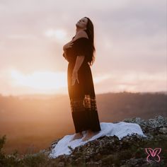 a woman in a black dress standing on top of a hill with the sun behind her