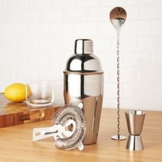 a stainless steel cocktail shaker, measuring spoon and strainer on a wooden table