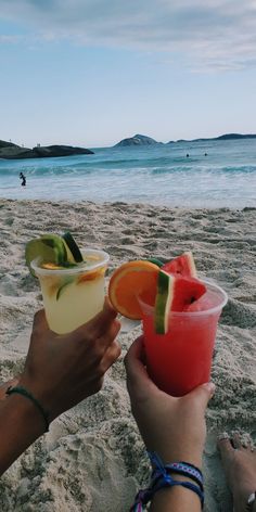 two people holding drinks on the beach with watermelon and avocado slices