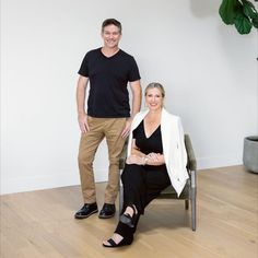 a man standing next to a woman sitting on a chair in front of a white wall