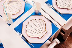 the table is set with pink and blue place mats, plates, silverware, and seashells