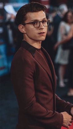 a man wearing glasses standing in front of a red carpet with people looking on from behind him