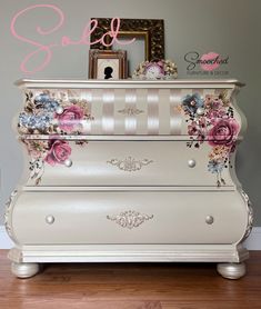 a white dresser with flowers painted on the top and bottom, sitting in front of a wall