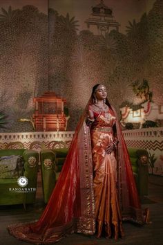 a woman in a red and gold sari standing next to a green couch with an ornate wallpaper behind her