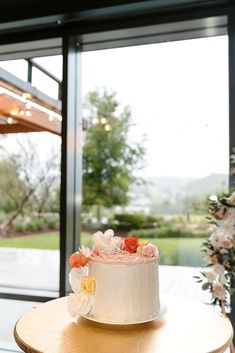 a white cake sitting on top of a wooden table next to a glass wall with flowers