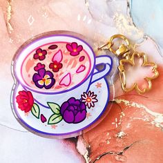 a keychain with a coffee cup and flowers painted on it, sitting next to a flowered background