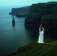 a woman standing on top of a lush green hillside next to the ocean with cliffs in the background