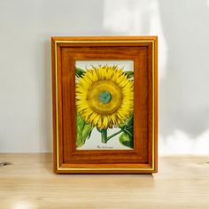 a painting of a yellow sunflower in a wooden frame on a table with a white wall behind it