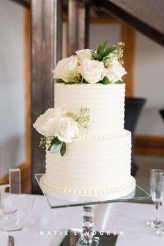 a wedding cake with white flowers and greenery sits on a glass stand at the head table