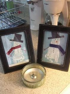 two framed snowmen sitting on top of a counter next to a cup and saucer