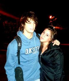 a young man and woman standing next to each other at night with lights in the background