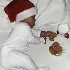 a baby in a santa hat eating cookies and milk from a bowl on a bed