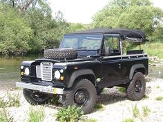 an old black land rover is parked on the side of a river bank with trees in the background